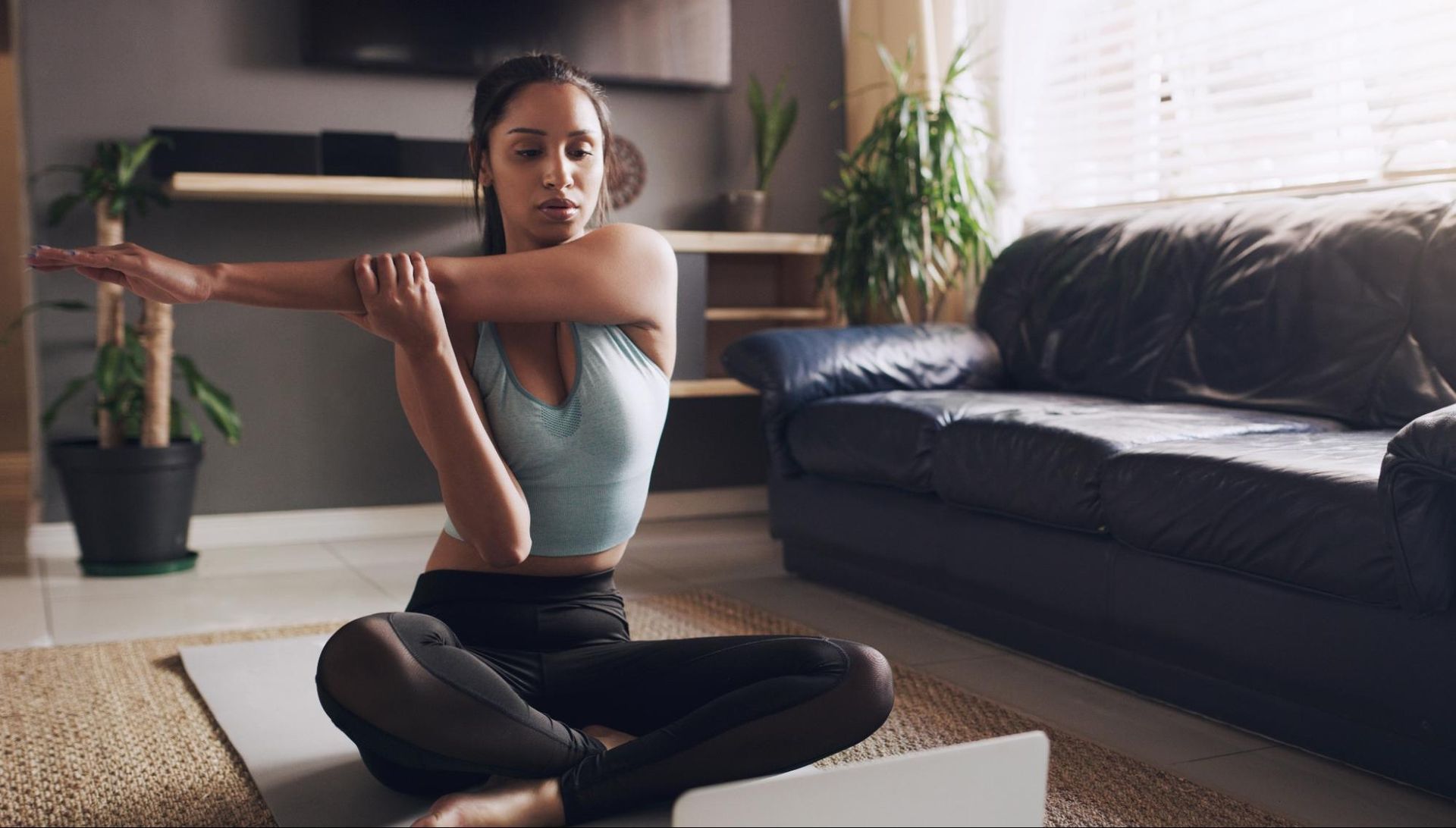 A woman stretches on a yoga mat next to a couch and potted plant.