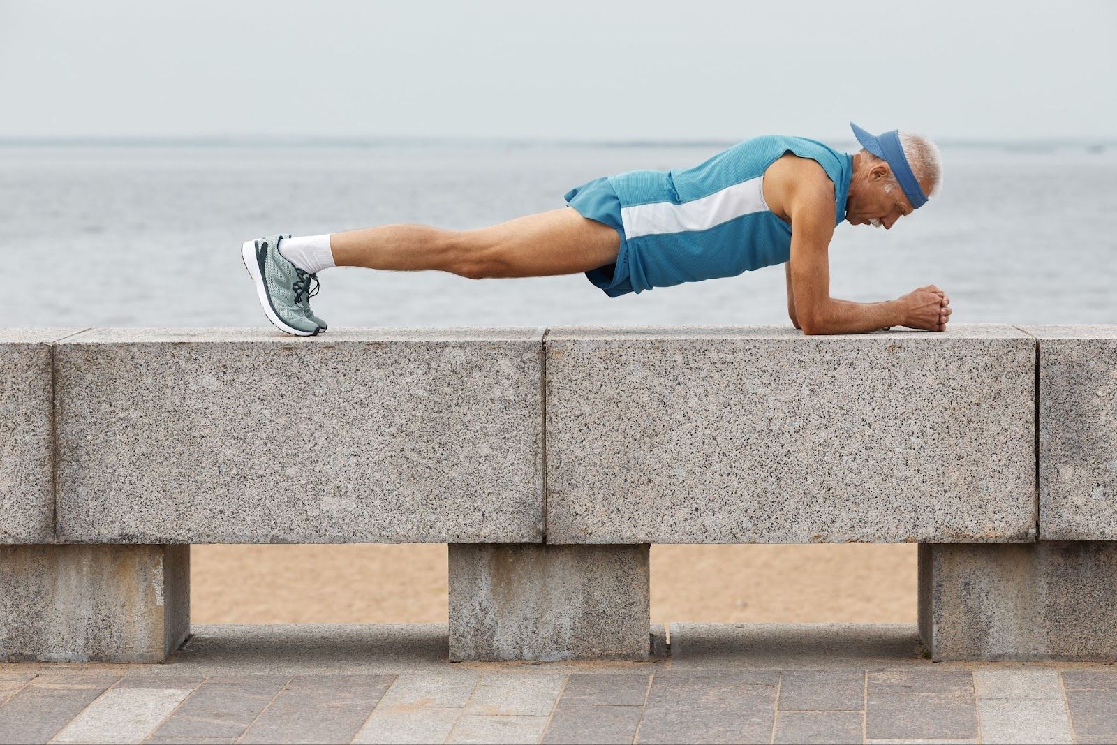 A senior sports enthusiast wearing a blue and white jersey and shorts does a plank exercise on a ledge by the bay.
