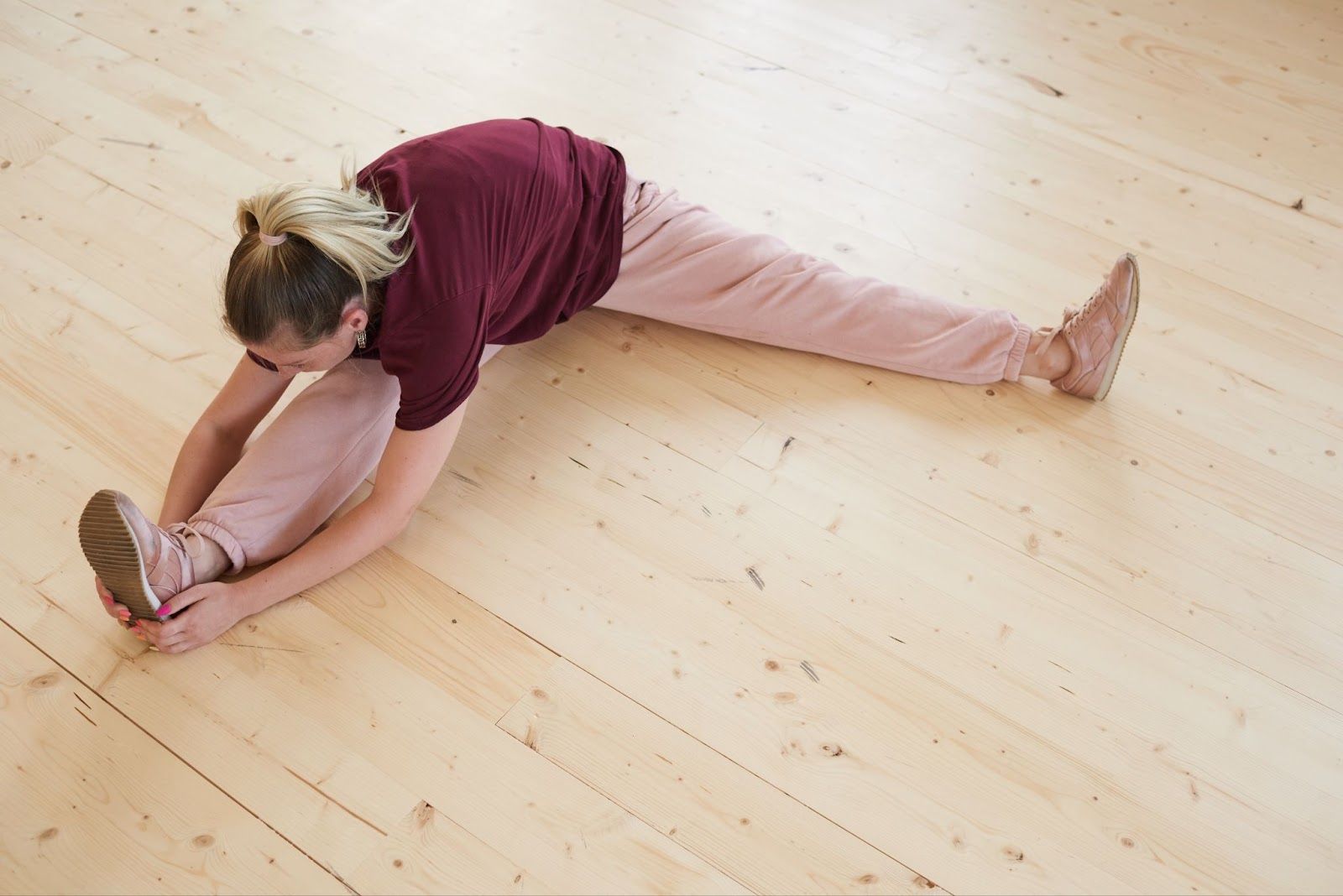 A young and healthy woman wearing red-pink sporty attire is sitting on the floor and doing the stretching.
