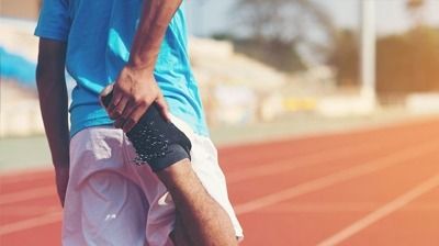 Athlete stretching leg on a red track; wearing blue shirt and white shorts. Sunny outdoor setting.