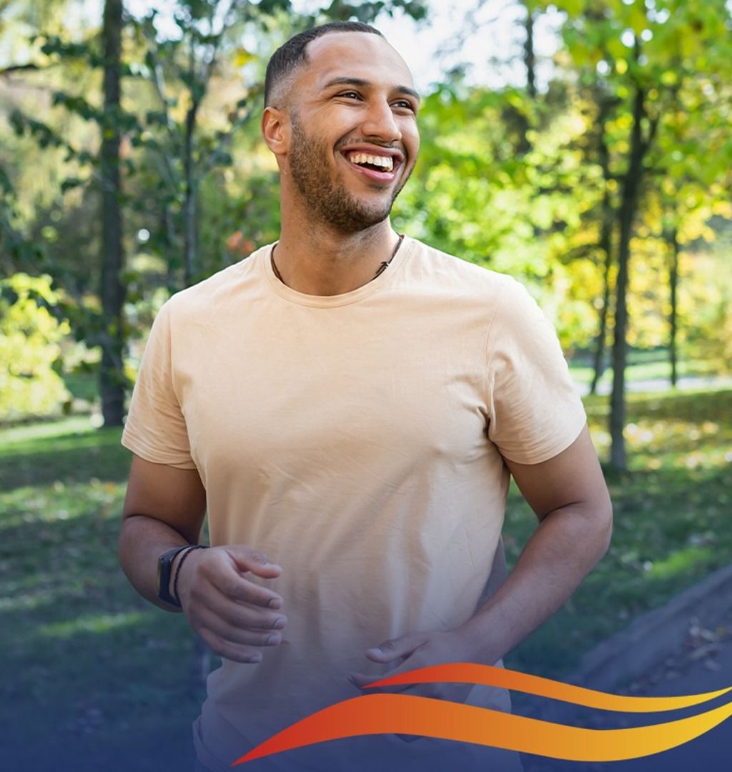 Man smiling while jogging outdoors. Wearing a tan t-shirt, trees in the background.