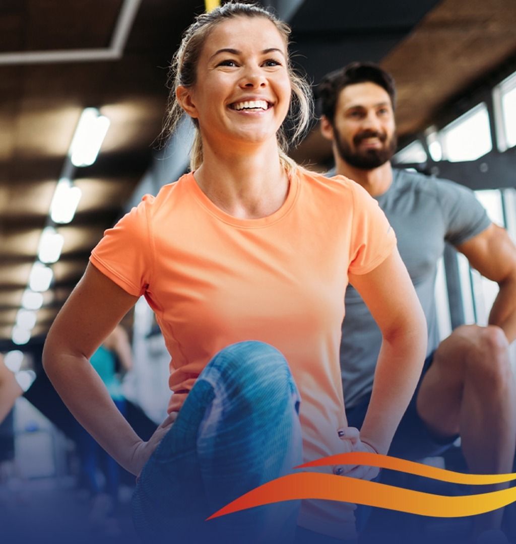 Woman in orange shirt smiles while stretching in a gym with a man.