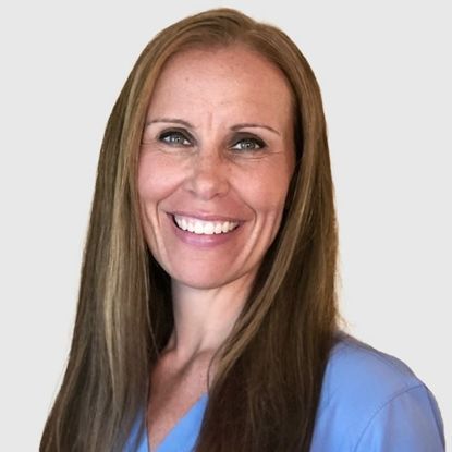 Woman with long, light brown hair smiles, wearing blue scrubs.