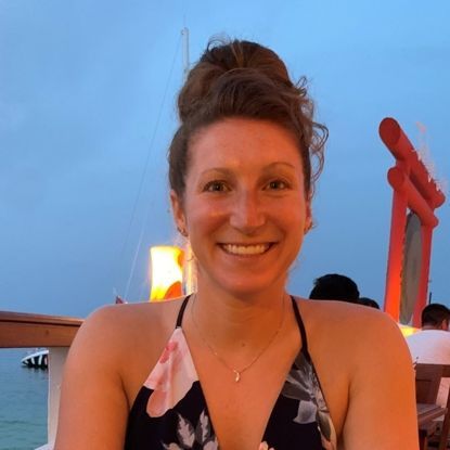 Woman with bun smiling, in floral top, near water at dusk.