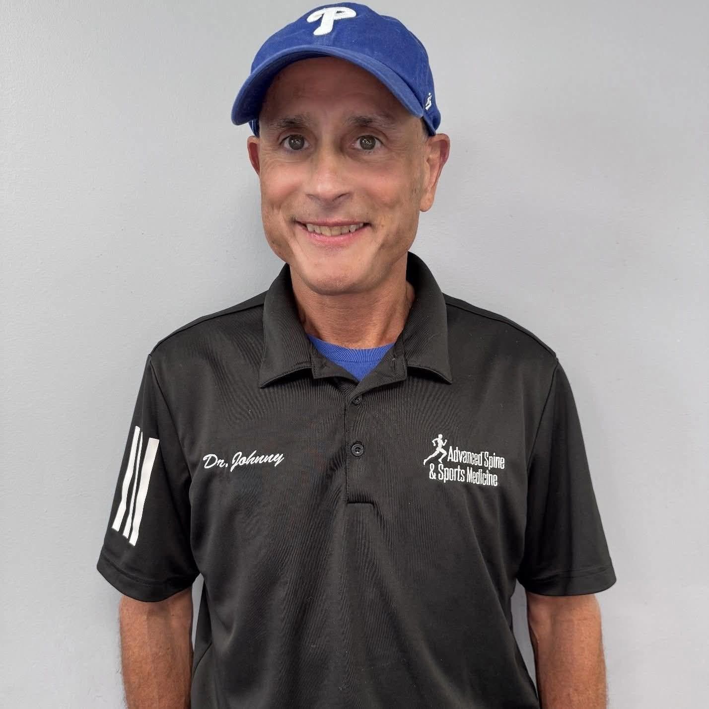 Man wearing a blue cap and black scrubs smiles in front of a gray wall.