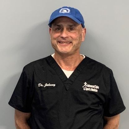 Man wearing a blue cap and black scrubs smiles in front of a gray wall.