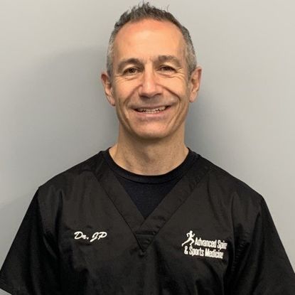 Man in black scrubs with a logo, smiling, stands in front of a gray wall.