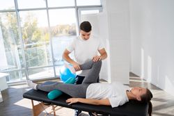 Physical therapist stretching patient's leg on a treatment table; sunny room with large window.