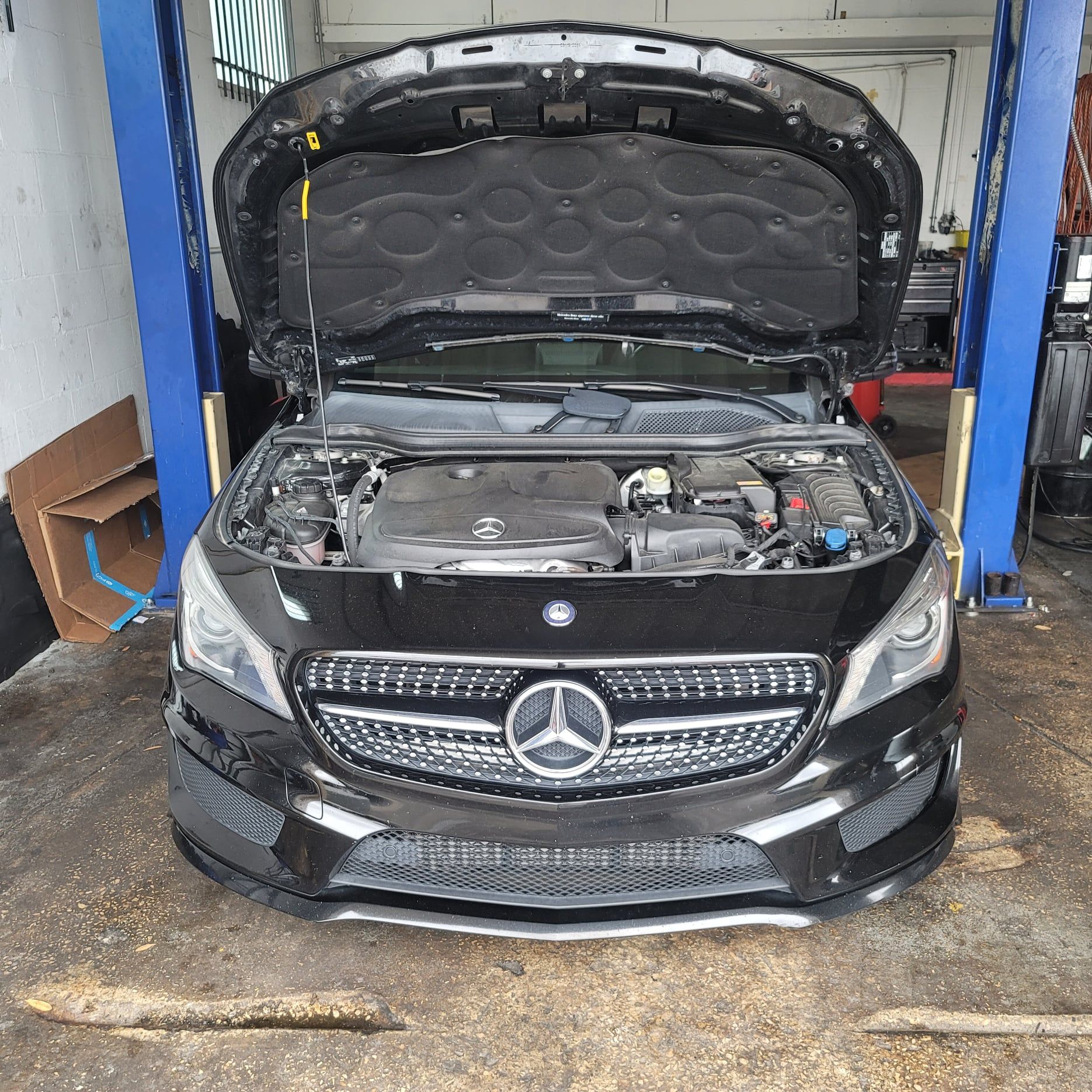 A black mercedes benz with the hood up is on a lift in a garage.