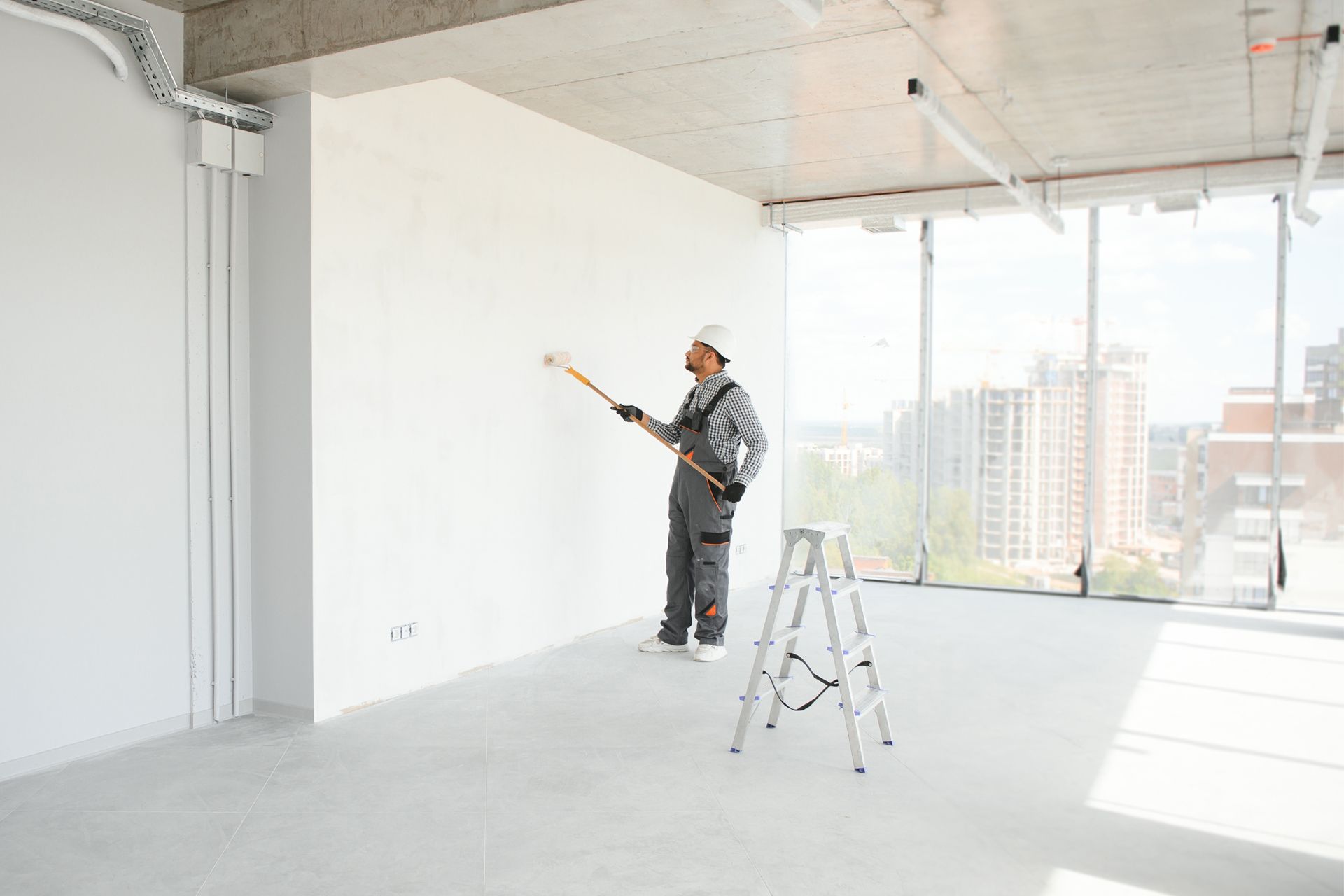 A worker in gray overalls paints a white wall in a bright, empty room, with a ladder and city view.