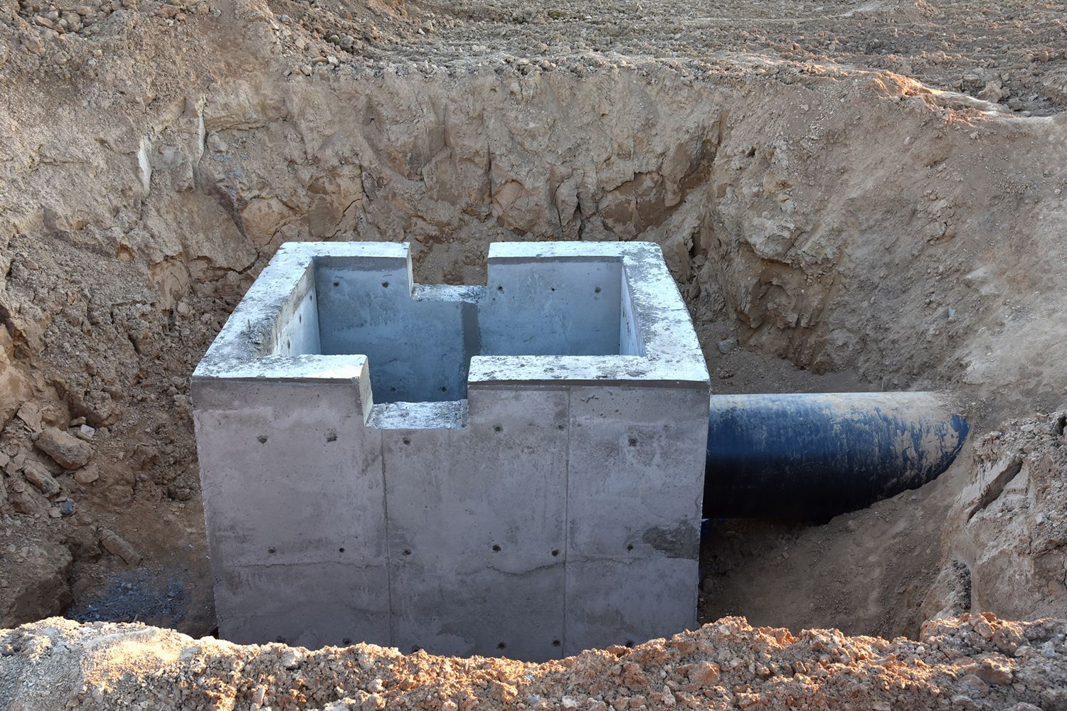 Concrete drainage structure in an earthen excavation, with a black pipe extending from its side.