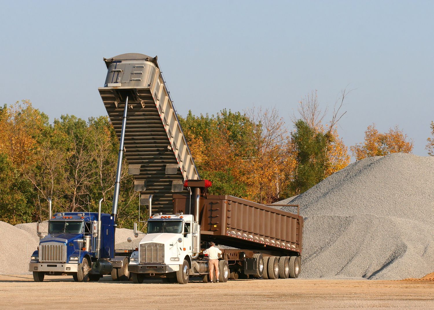 Two dump trucks unloading gravel at a quarry; a man stands near.