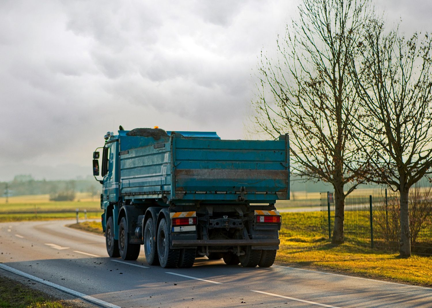 Blue dump truck on a road, driving away, with fields and trees in the background under a cloudy sky.