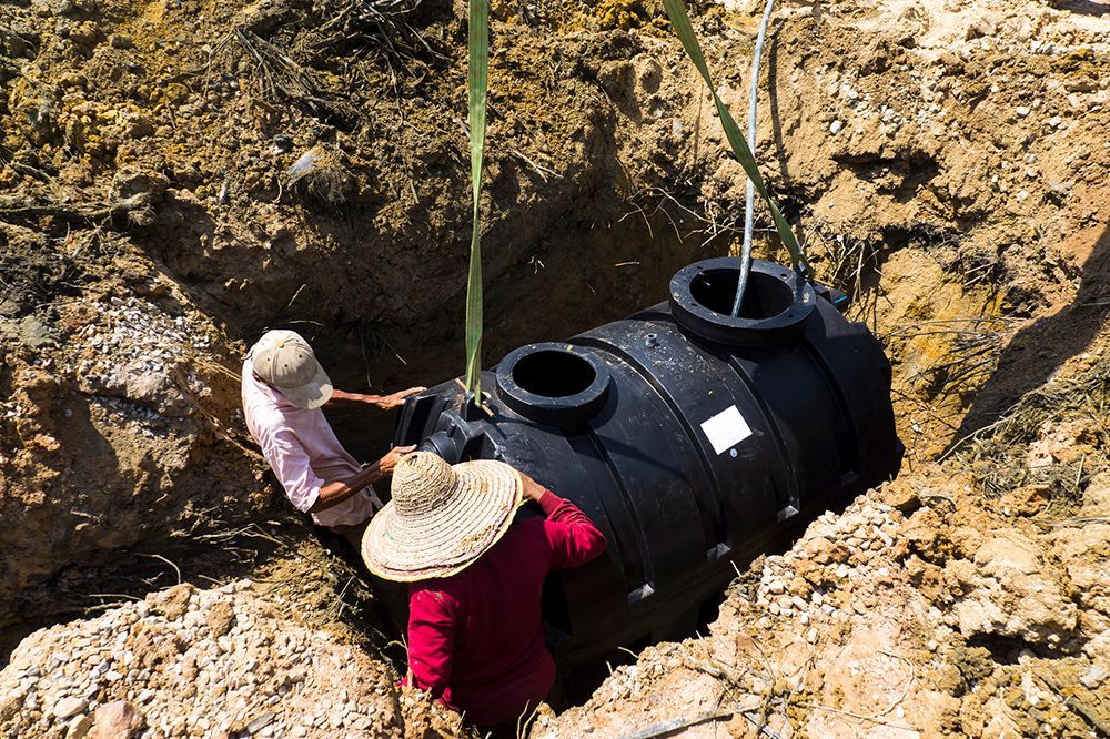 Two workers installing a black septic tank into a hole in the ground.