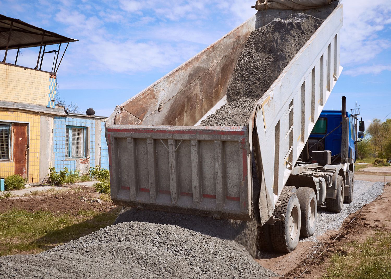 Dump truck unloading gravel onto a road near a small building on a sunny day.