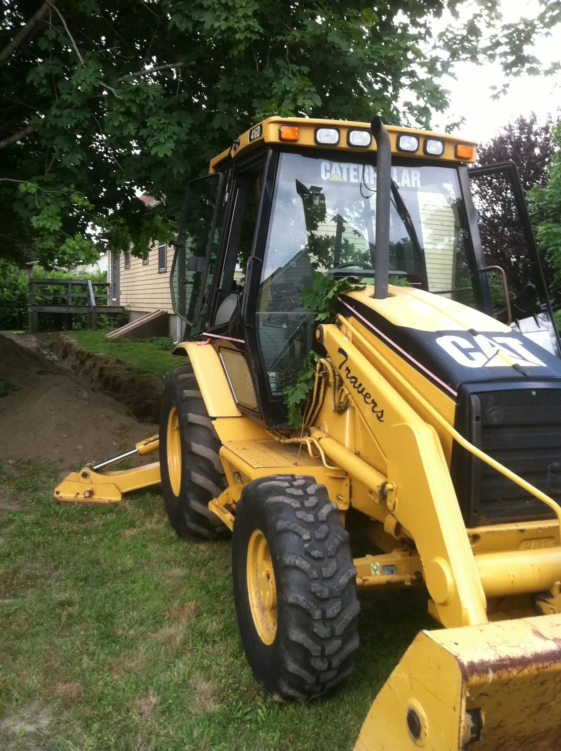 Yellow Caterpillar backhoe digging in a yard, near a house and trees.