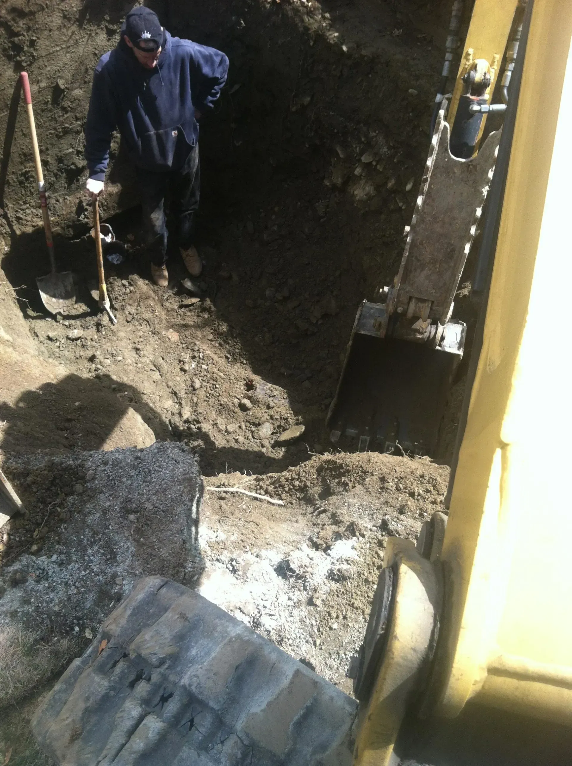Person standing in a trench, alongside an excavator. The person is holding a shovel. Sunny, outdoor setting. Person standing in a trench, alongside an excavator. The person is holding a shovel. Sunny, outdoor setting.