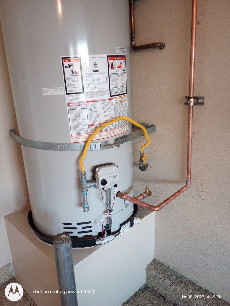 A water heater with gas line and copper pipes against a tan wall in a garage.