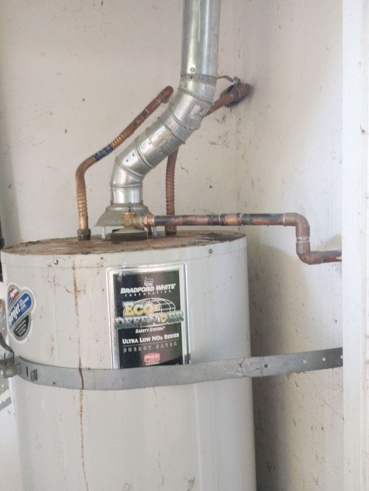 Water heater with rusted pipes and vent in a corner against a white wall.