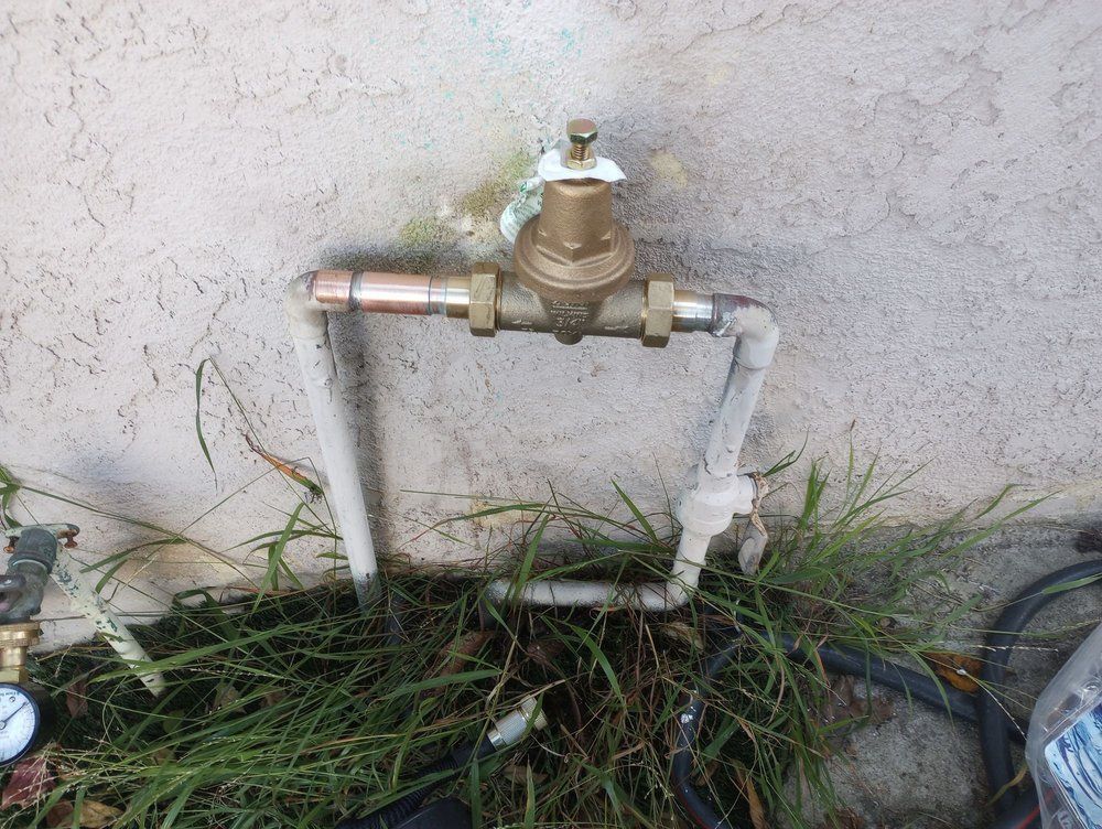 Outdoor water shut-off valve with copper and PVC piping. The valve is on a concrete wall surrounded by overgrown grass.
