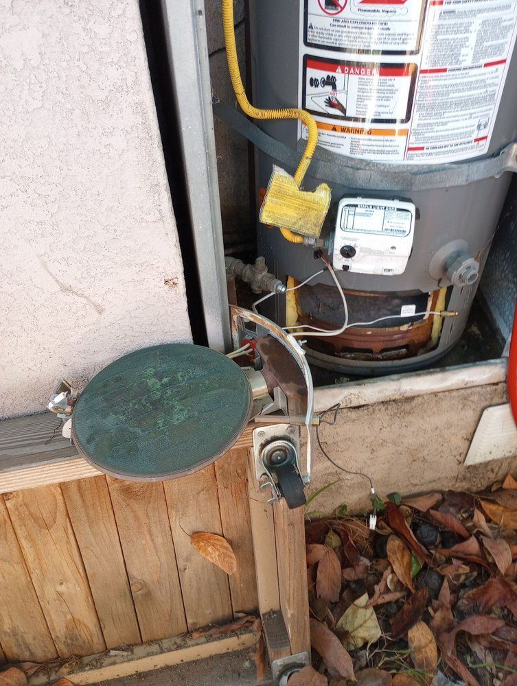 Water heater with exposed wiring next to a round, green metal cover and wooden siding.
