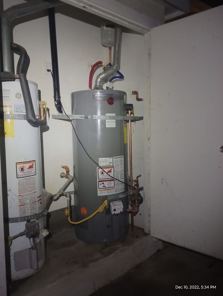 Two water heaters, one gray and one white, in a utility closet with pipes and a door.