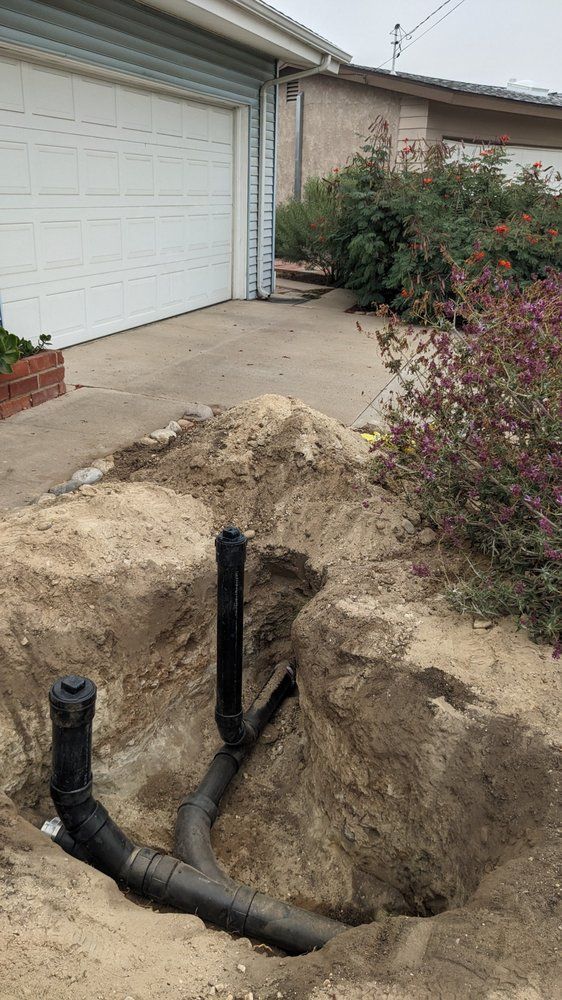 Excavated ground with exposed black plumbing pipes near a garage and house.