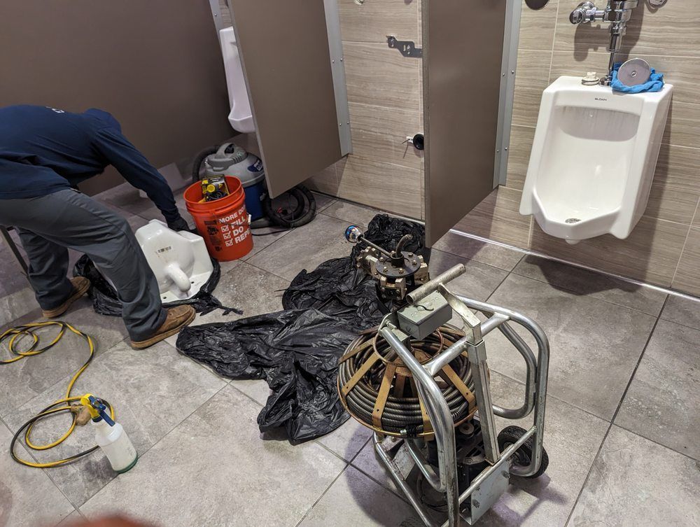 Plumber working in a public restroom. He's near a urinal and toilet stall, with tools and a snake on the floor.