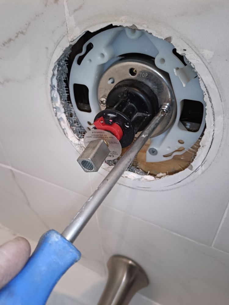 A hand using a screwdriver to work on a faucet cartridge inside a tiled shower.