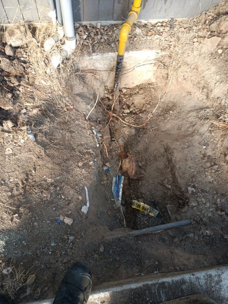 Trench dug near a sidewalk, revealing underground pipes. Yellow gas line visible.