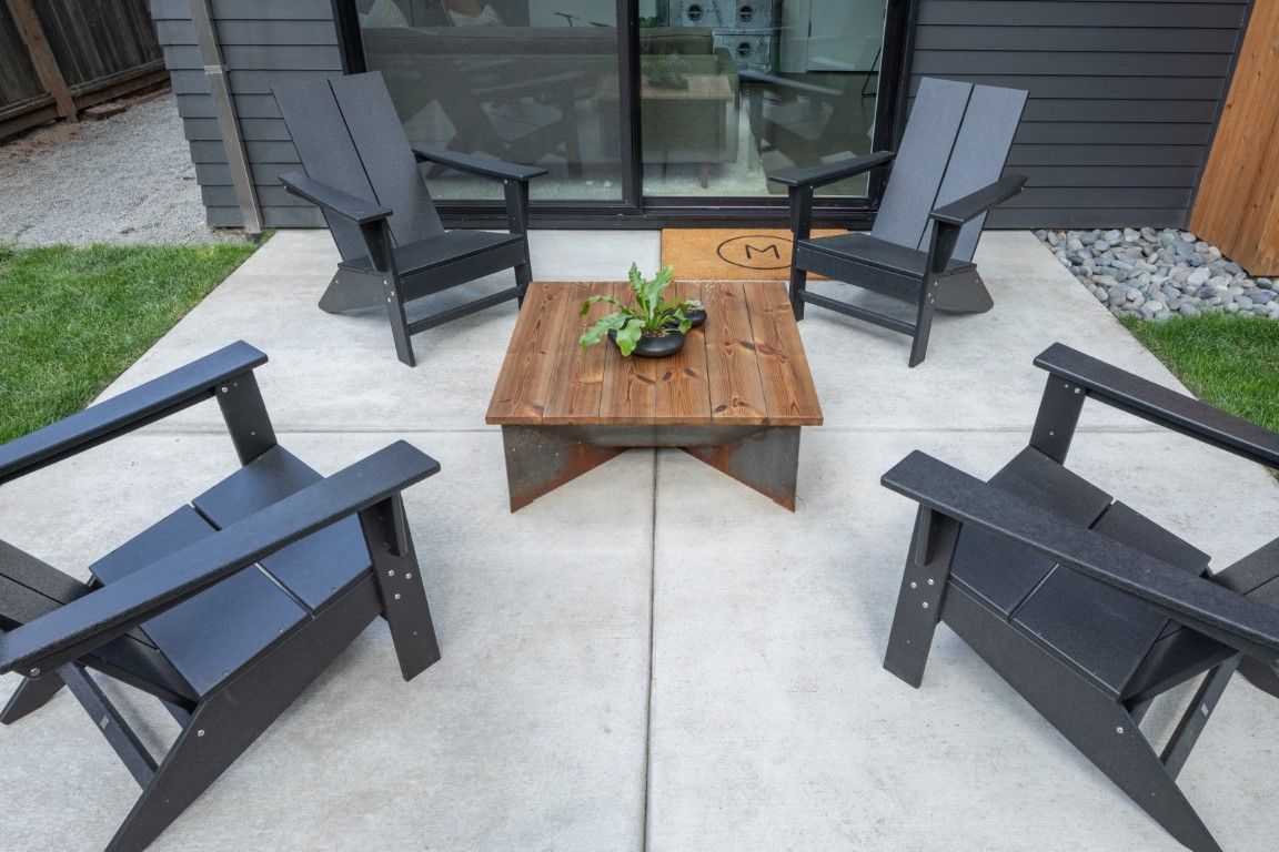 Four black Adirondack chairs surround a wooden square coffee table with greenery on a concrete patio.