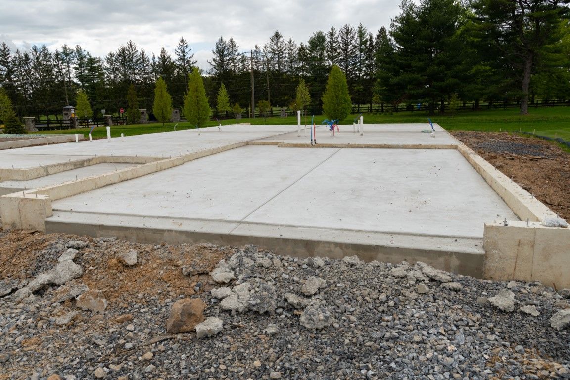 Concrete foundation of a building under construction. Grey concrete, dirt, and trees in the background.