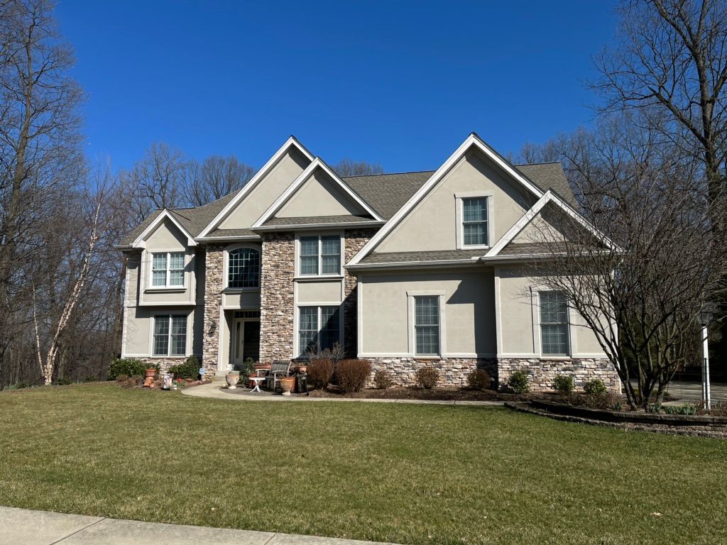 A beige, two-story house with stone veneer accents sits on a grass lawn under a clear blue sky, surrounded by bare trees.