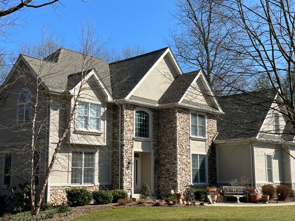 A large, two-story house with stone and light-colored siding, a dark shingled roof, and green trees against a blue sky.