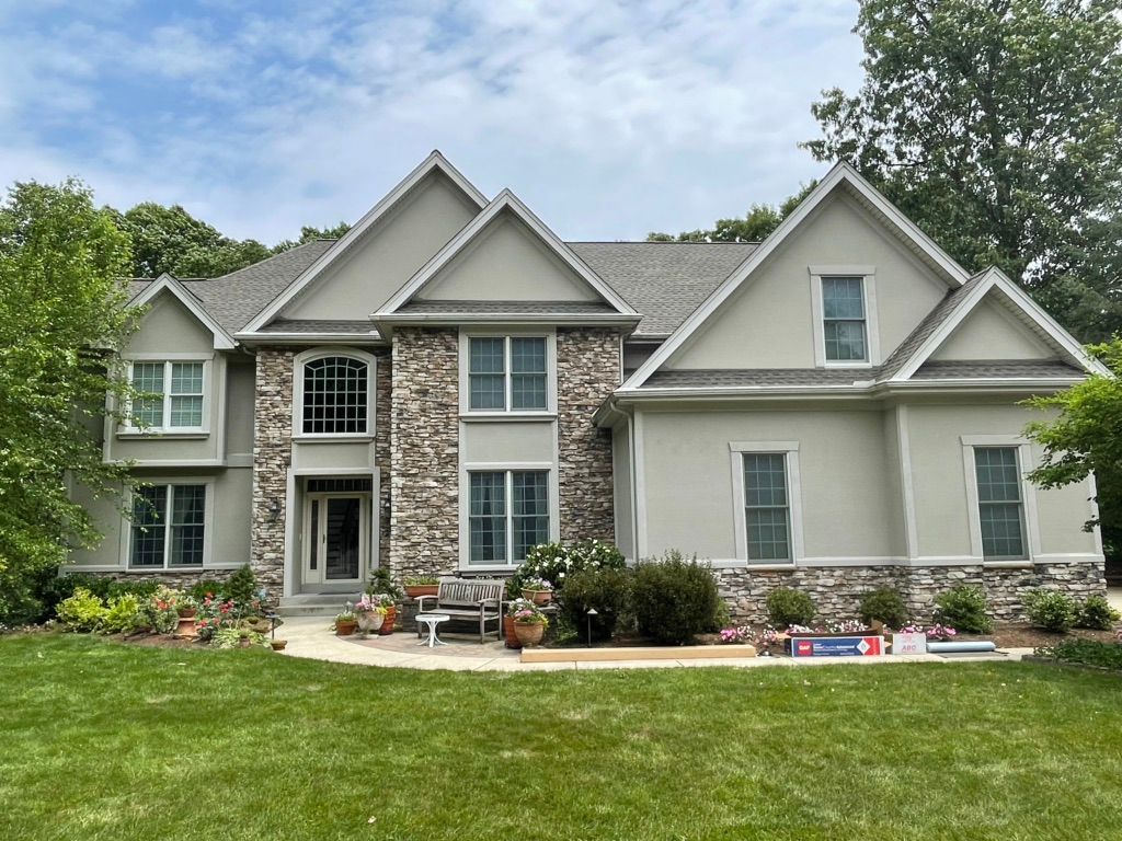 A two-story house with a light gray stucco exterior, stone facade accents, dark shingled roof, and green lawn.