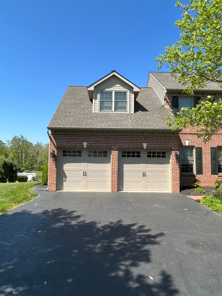 A two-story brick suburban house with a two-car garage, a dormer window above, and a paved driveway on a sunny day.
