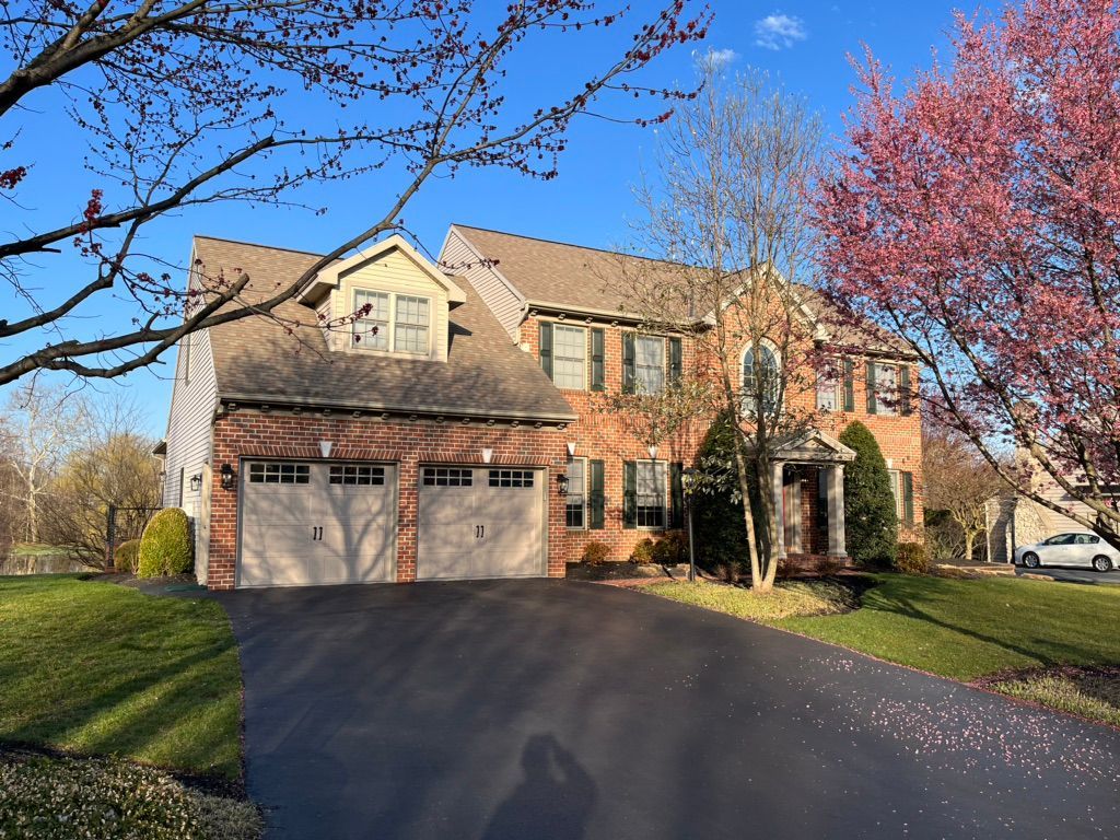 A two-story brick suburban house with a two-car garage, a paved driveway, and flowering trees under a clear blue sky.
