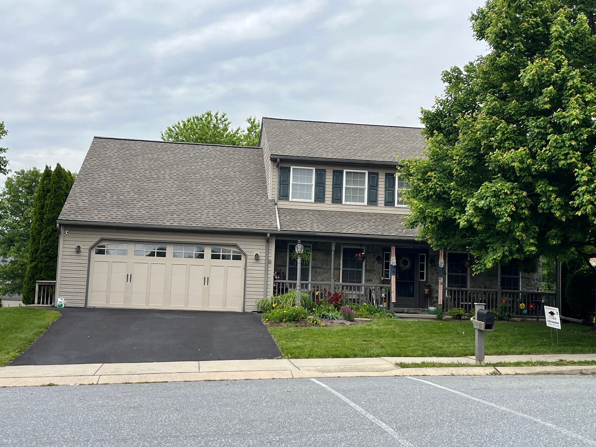 A two-story stone house with a side-attached garage, dark shingled roof, and a mature tree in the front yard.