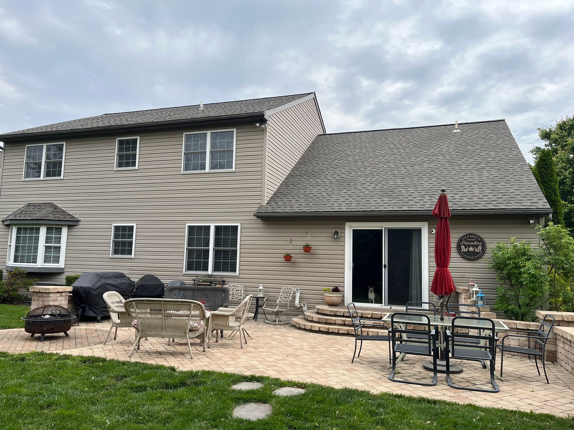 A two-story tan house with a stone patio, outdoor furniture, a red umbrella, and a grassy backyard under a cloudy sky.