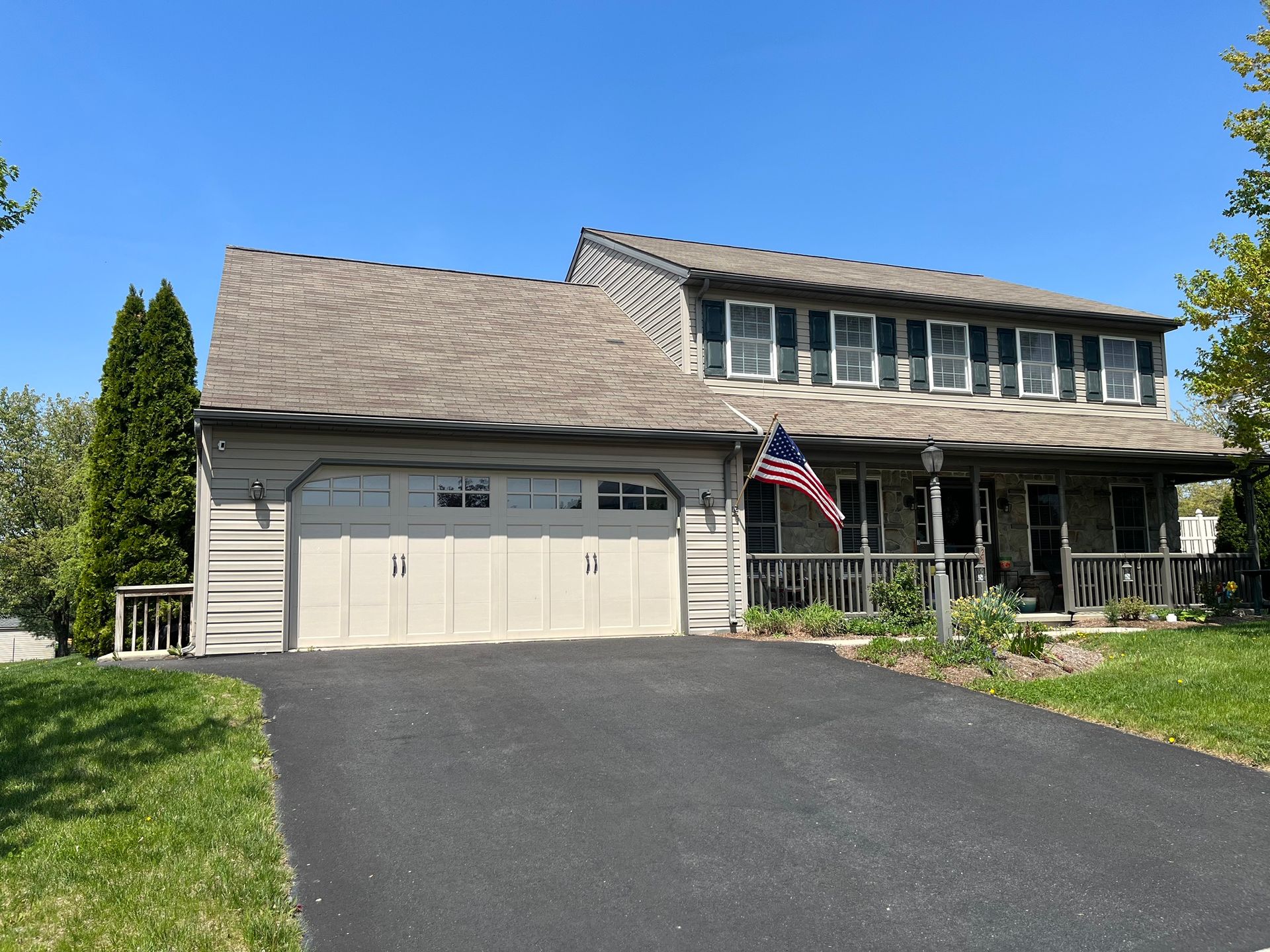 A two-story, light-colored suburban house with a two-car garage, a front porch, and an American flag on a sunny day.