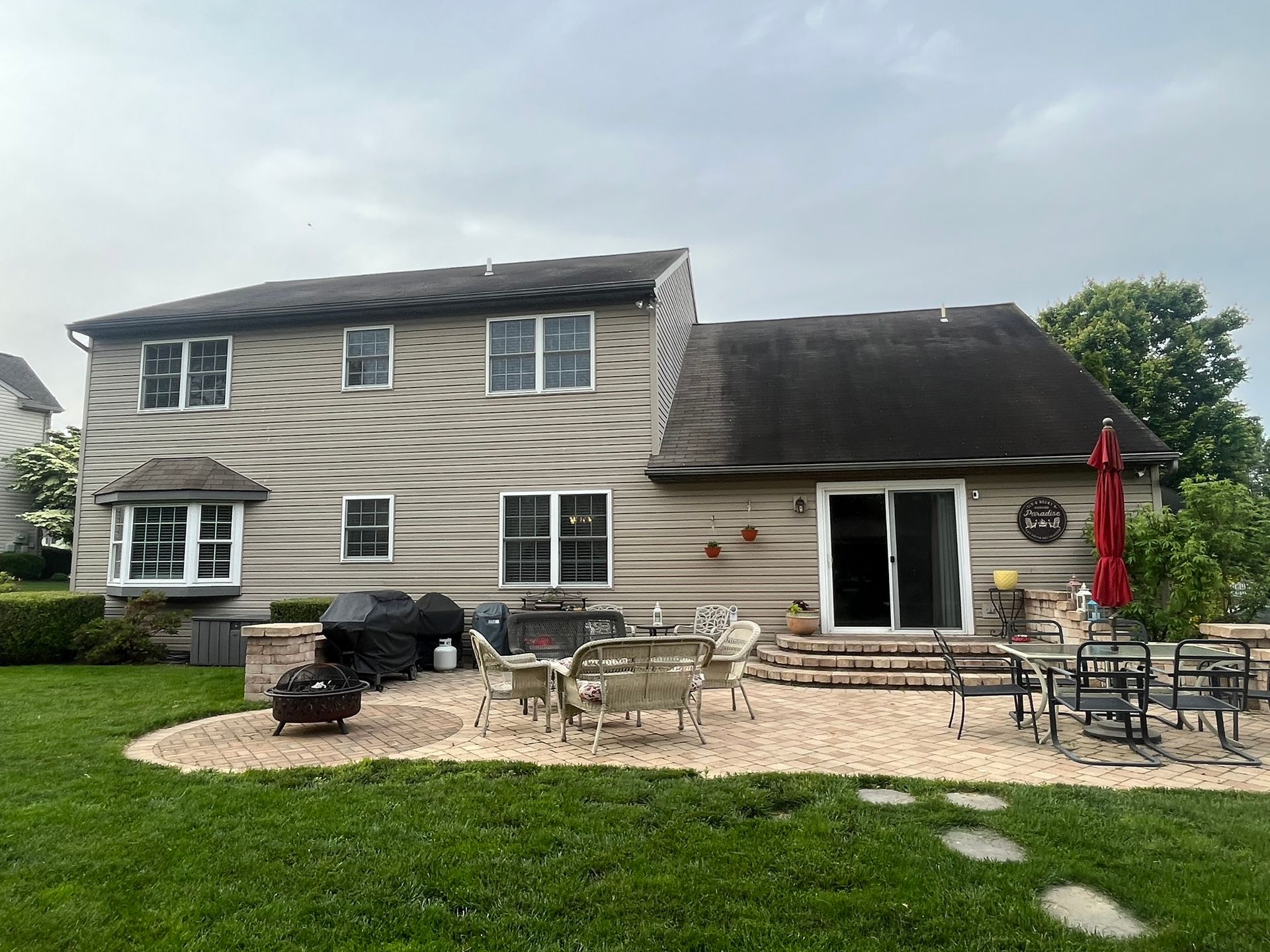 A two-story tan house with a stone patio, outdoor furniture, a grill, and a green lawn under a cloudy sky.