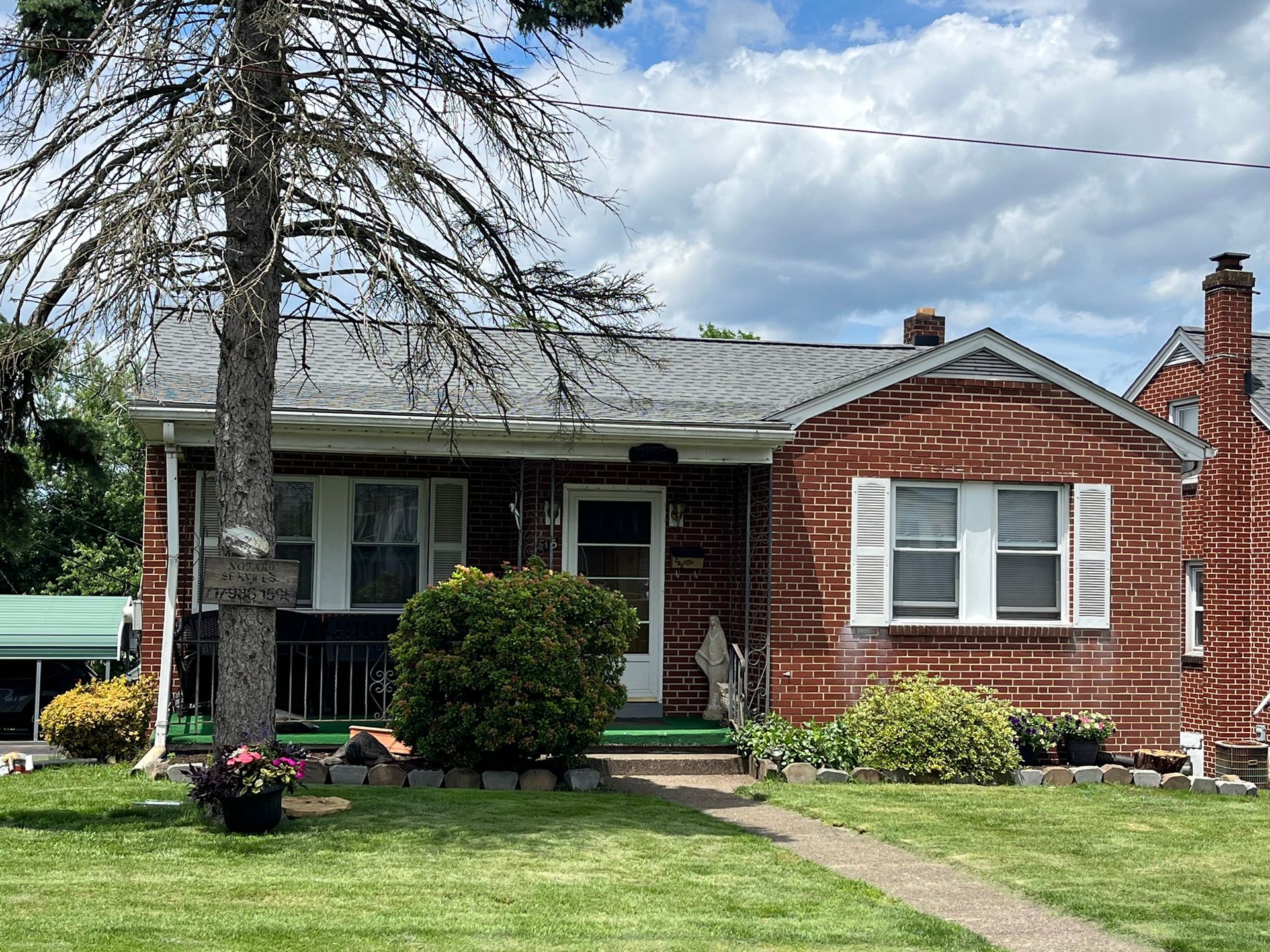 A single-story red brick house with a white front door, shuttered windows, and a green porch under a cloudy sky.