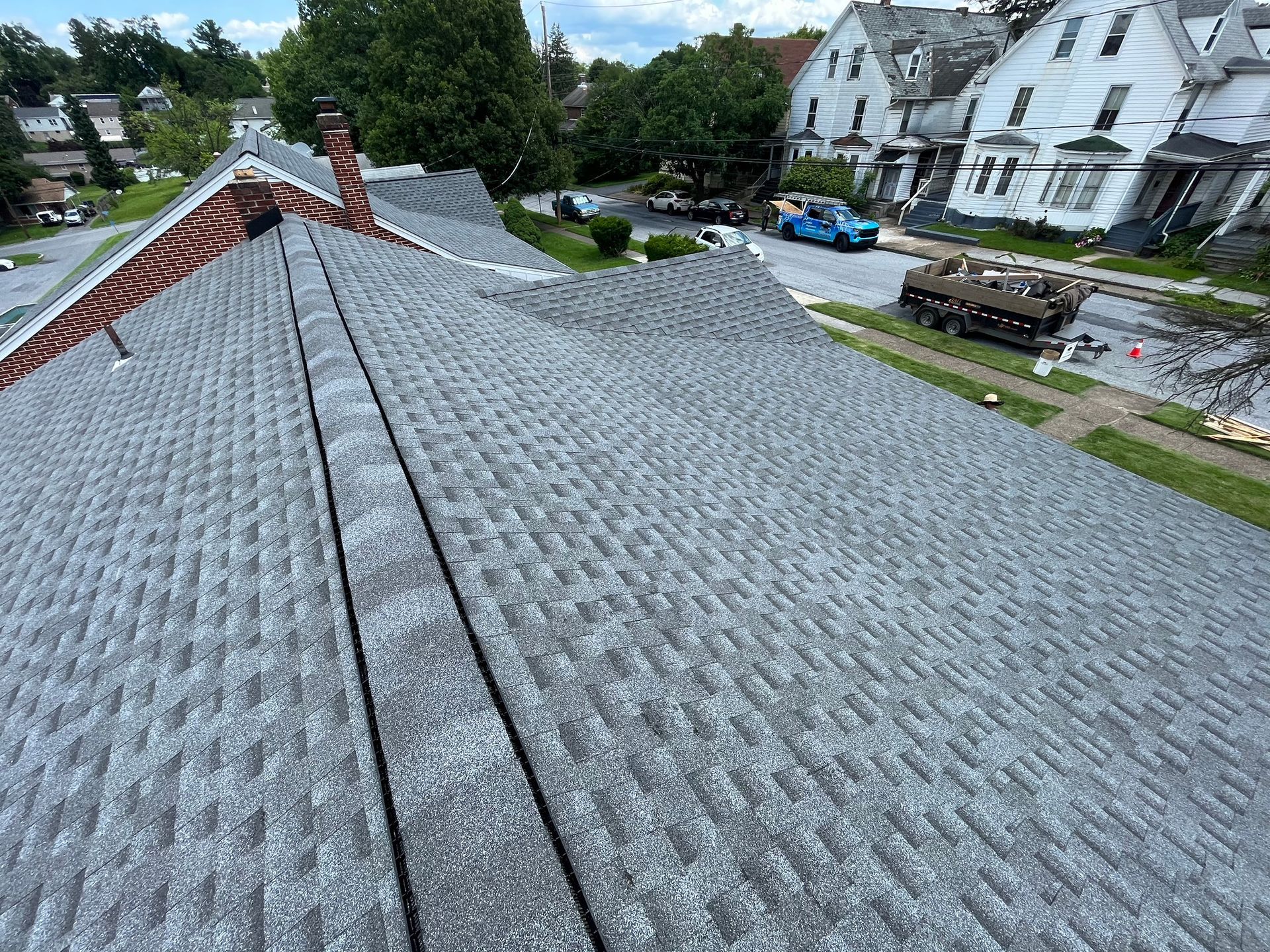 A high-angle view looking down at a gray asphalt shingle roof on a sunny day, with houses and a street visible below.