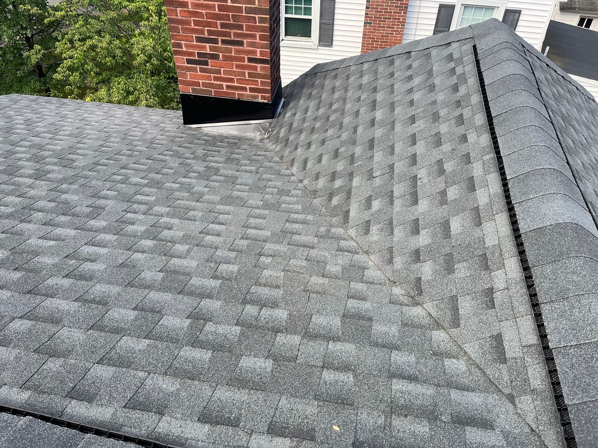 A high-angle view of a gray shingled roof with a red brick chimney and a prominent roof valley.