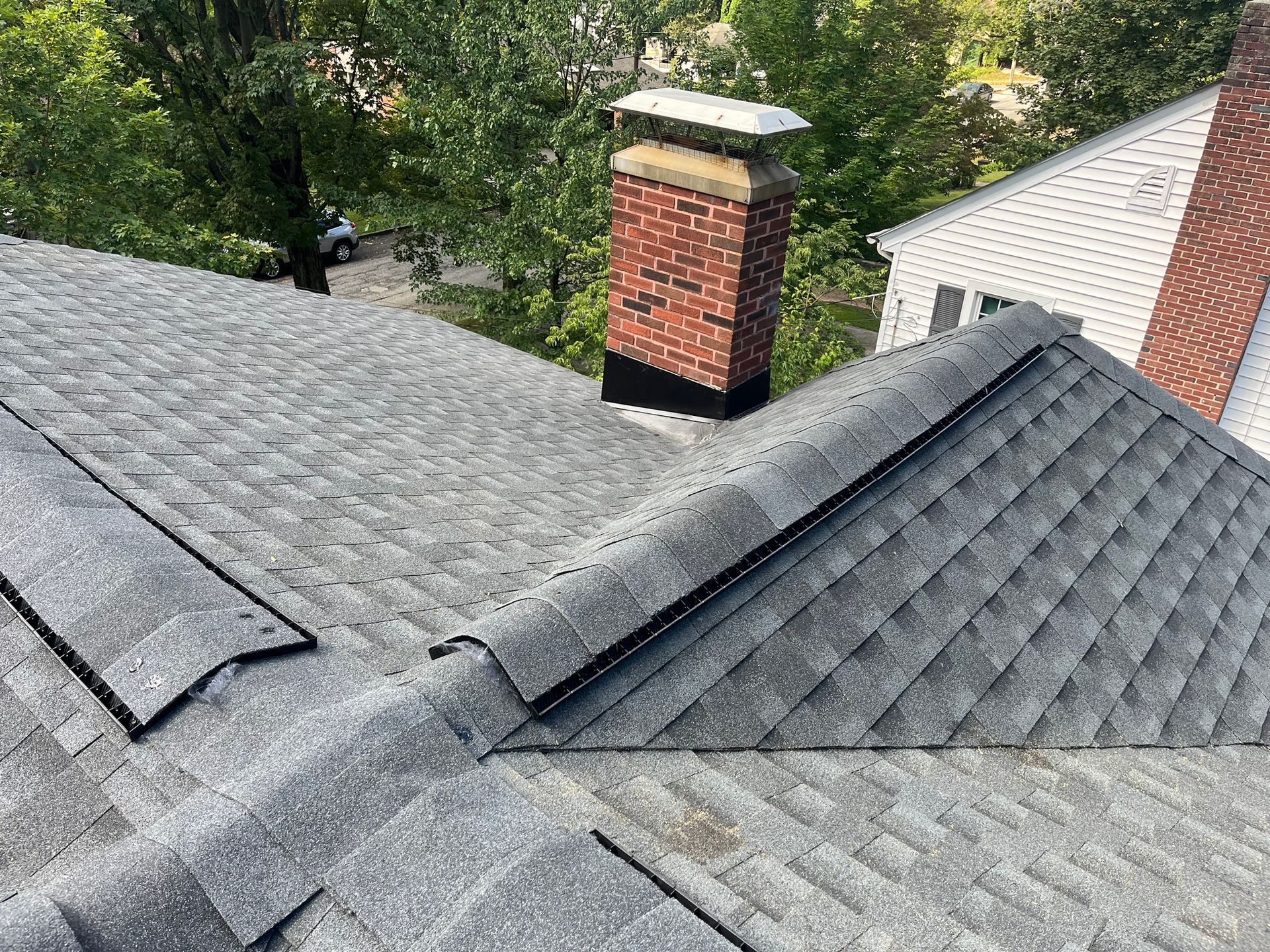 A high-angle view of a gray shingled roof with a red brick chimney and a nearby house roof in a suburban setting.