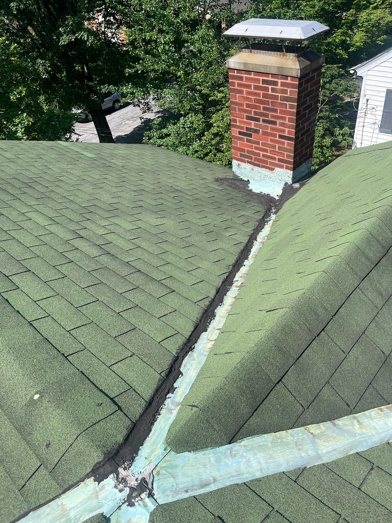 A green shingled roof with a brick chimney, a dark valley flashing, and a light-colored metal ridge cap.