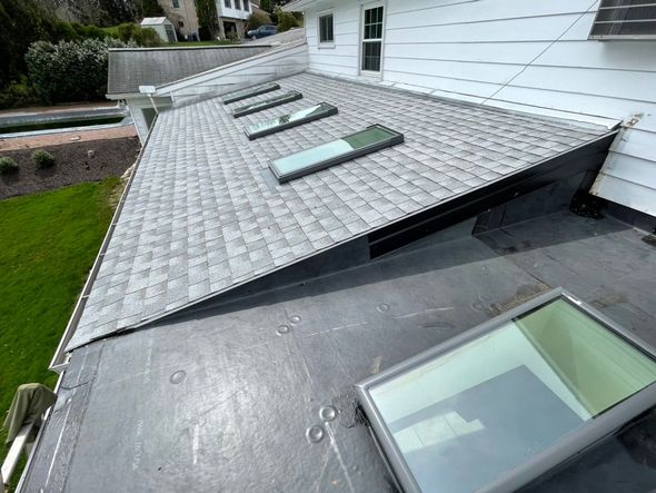 A high-angle view of a grey shingled roof with three skylights, adjoining a flat roof with one large skylight.
