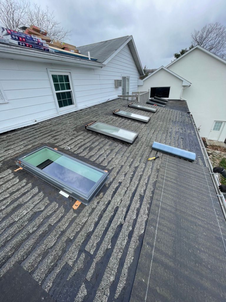 A flat roof with four rectangular skylights, featuring worn, textured roofing material and adjacent white house siding.