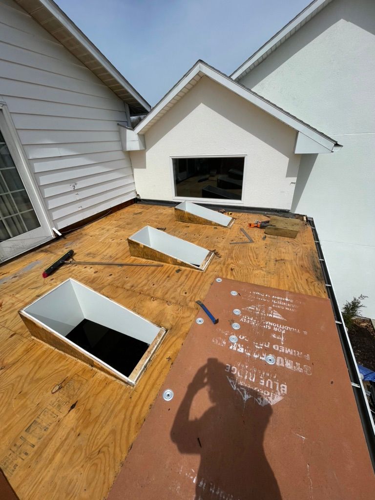 A flat, plywood-covered roof under construction featuring three skylight openings and a wall with a large window.