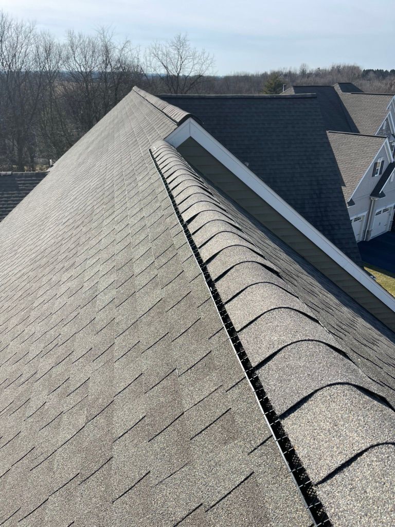 A high-angle view looking down the hip of a gray asphalt shingle roof toward a neighboring house under a blue sky.